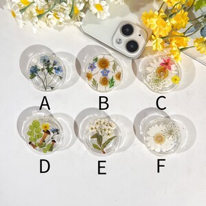 May include: Six round clear resin phone grips with different pressed flower designs. The designs include blue forget-me-nots, white daisies, a pink flower with white accents, green ferns and a brown mushroom, white flowers with green stems, and a white daisy with a brown centre.