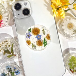 May include: A white phone case with a clear phone grip featuring pressed flowers. The flowers are white, yellow, and blue. The phone case has a white background and a black camera lens.