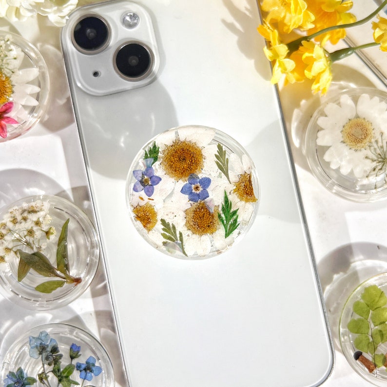 May include: A white phone case with a clear phone grip featuring pressed flowers. The flowers are white, yellow, and blue. The phone case has a white background and a black camera lens.