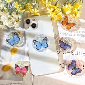 May include: Four phone grips with butterfly designs. The grips are clear with glitter and feature different colored butterflies. The butterflies are blue, purple, pink, and orange.