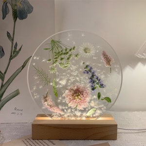 May include: A round, clear resin lamp base with pressed flowers and a wooden base. The lamp features a variety of dried flowers, including pink, purple, and white blooms.