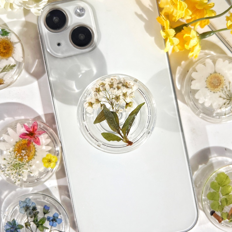 May include: A white phone case with a clear phone grip featuring a pressed white flower with green leaves.
