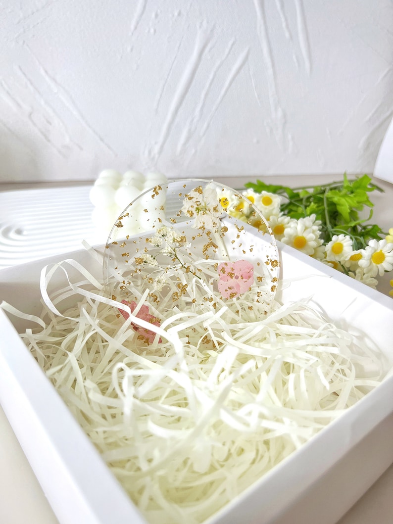 May include: A round, clear resin coaster with dried flowers and gold flakes. The coaster is in a white box with white shredded paper.