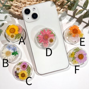May include: A collection of clear phone grips, each featuring a unique arrangement of dried flowers in various colors. The grips are attached to the back of a white smartphone, showcasing the floral designs. The flowers include yellow, pink, orange, purple, and white.