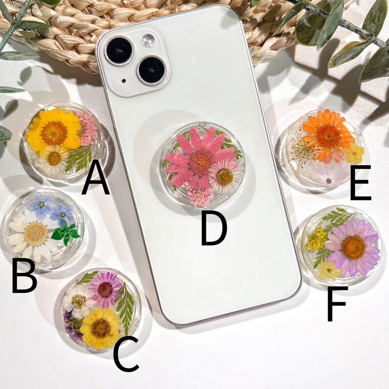 May include: A collection of clear phone grips, each featuring a unique arrangement of dried flowers in various colors. The grips are attached to the back of a white smartphone, showcasing the floral designs. The flowers include yellow, pink, orange, purple, and white.
