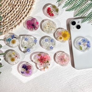 Puede incluir: Surtido de 12 agarres para teléfono con varias flores y hojas prensadas encapsuladas en resina transparente. Los agarres para teléfono son redondos y tienen una parte posterior plana para una fácil fijación a una funda de teléfono.