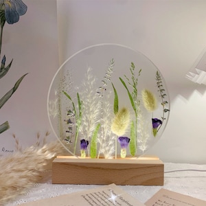 May include: A round, clear glass lamp base with a wooden stand. The glass base contains pressed flowers and grasses in shades of green, white, and purple.