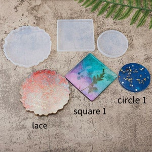 May include: Four silicone molds for resin crafts. The molds are in the shapes of a circle, square, and two different lace designs. The molds are on a textured gray surface.