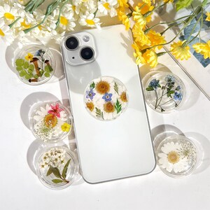 May include: A white iPhone with a floral phone grip. The phone grip is a clear circle with a pressed flower design. The phone is surrounded by white and yellow flowers.