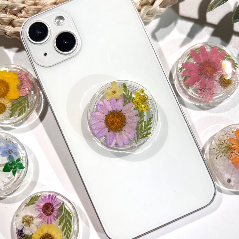 May include: Clear, round phone grips with dried flowers in various colors, including yellow, pink, purple, and orange, are displayed on a white phone. The grips are attached to the back of the phone and are surrounded by other loose grips.