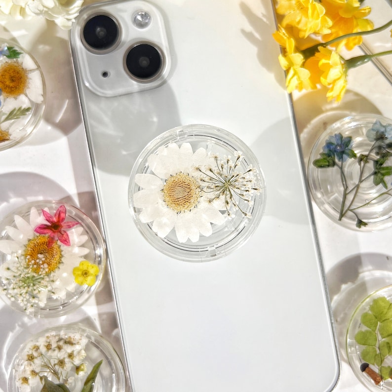 May include: A clear phone grip with a white daisy and a small green stem inside. The grip is on a white phone case with a camera lens.