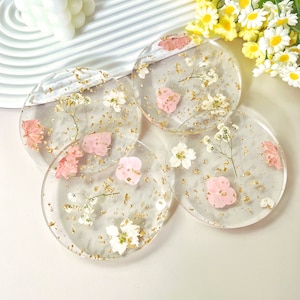 May include: Four round clear resin coasters with dried pink and white flowers and gold flecks embedded in the resin.