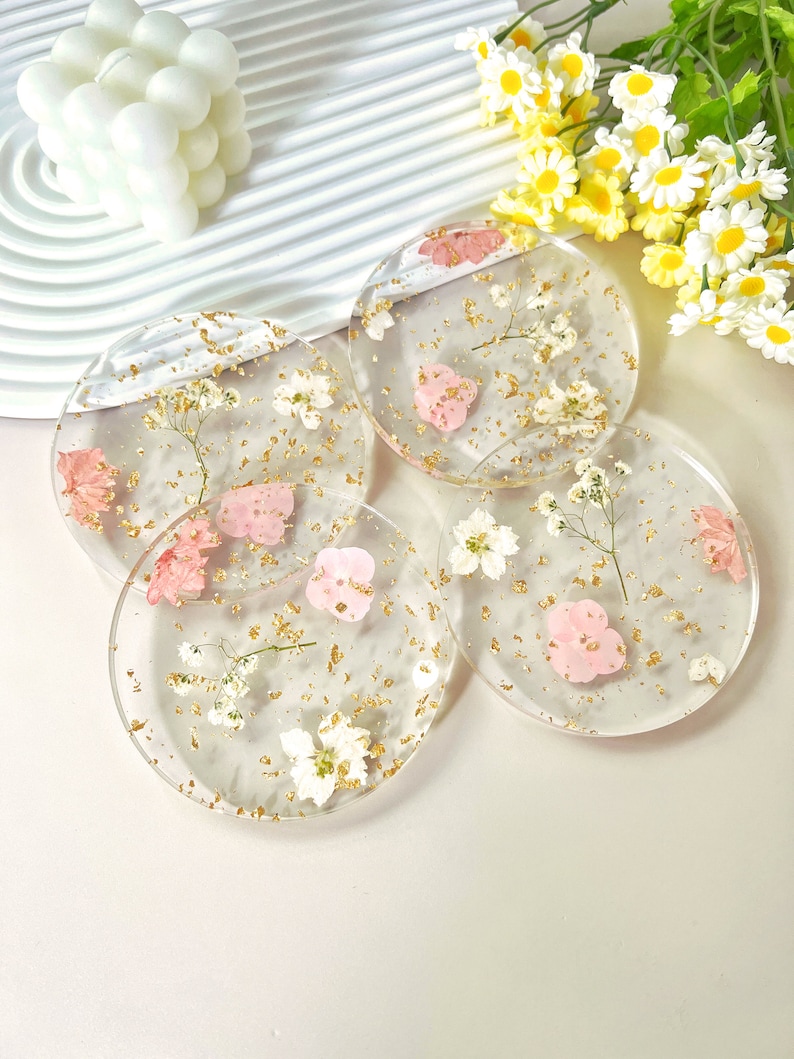 May include: Four round clear resin coasters with dried pink and white flowers and gold flecks embedded in the resin.