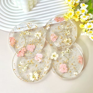 May include: Four round clear resin coasters with dried pink and white flowers and gold flecks embedded in the resin.