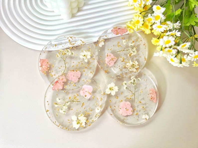 May include: Four round clear resin coasters with dried pink and white flowers and gold flecks embedded in the resin.