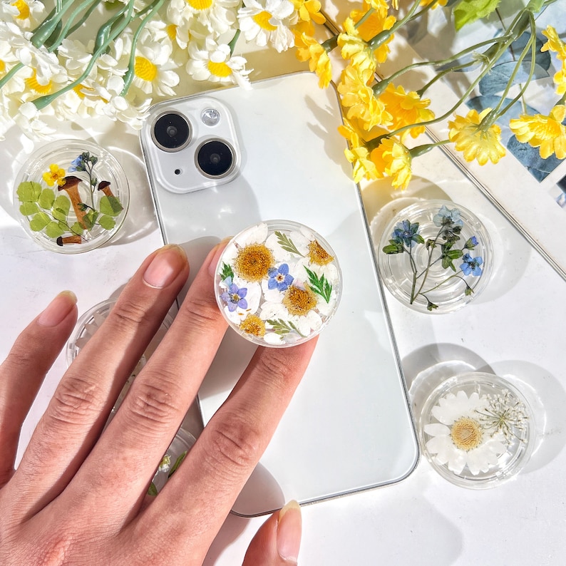 May include: A white iPhone with a clear phone grip featuring pressed flowers. The phone grip is a popular accessory for smartphones and is designed to provide a secure hold on the device.