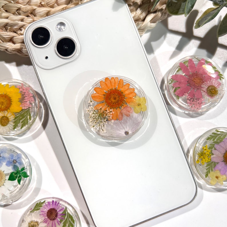May include: A white smartphone with a clear phone grip featuring an orange flower and other dried flowers. Several other floral phone grips with different colored flowers are visible.