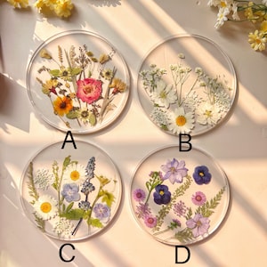 May include: Four round resin coasters with pressed flowers. The coasters feature different floral arrangements, including white, pink, blue, and purple flowers.