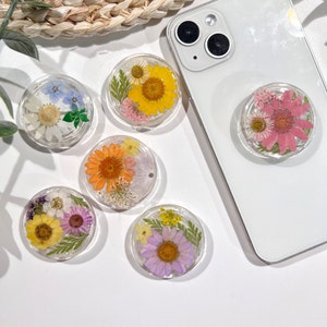 May include: A selection of clear, circular phone grips, each containing dried flowers set in resin. The grips showcase diverse flower varieties in yellow, orange, pink, purple, and white. One grip is affixed to a white smartphone.