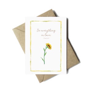 May include: A white greeting card featuring a watercolour sunflower illustration and the text "Do everything in love." The card has a yellow border and is paired with a brown envelope.