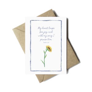 May include: A white greeting card with a blue border and a watercolour sunflower illustration. The card features the text "My heart leaps for joy, and with my song I praise Him. Psalm 28:7." The card is placed on a brown envelope.