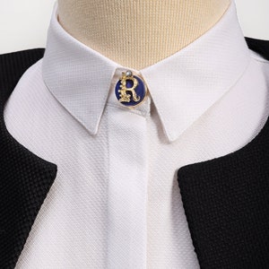 Peut inclure: Une chemise blanche à col avec une épingle bleue et dorée représentant la lettre "R" sur le col.