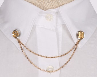 May include: A gold chain collar pin with two letter charms, one with the letter "S" and the other with the letter "H". The pin is attached to a white collared shirt.