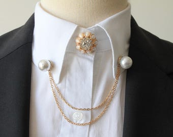 May include: A gold collar pin with a pearl and rhinestone design. The pin has a double chain attached to it, and the chain is also adorned with pearls.