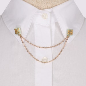 Puede incluir: Una camisa blanca con un broche de cadena de oro para el cuello con las letras 'E' y 'S' en oro.