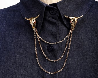 May include: A gold-tone collar pin with a skull design and a chain. The pin is attached to a black shirt.