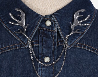 May include: A silver-toned collar pin with a chain and two antler-shaped pieces. The pin is attached to a denim shirt.
