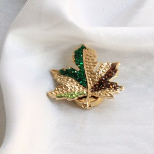 May include: A gold-toned brooch in the shape of a maple leaf with green, brown, and light green rhinestones.
