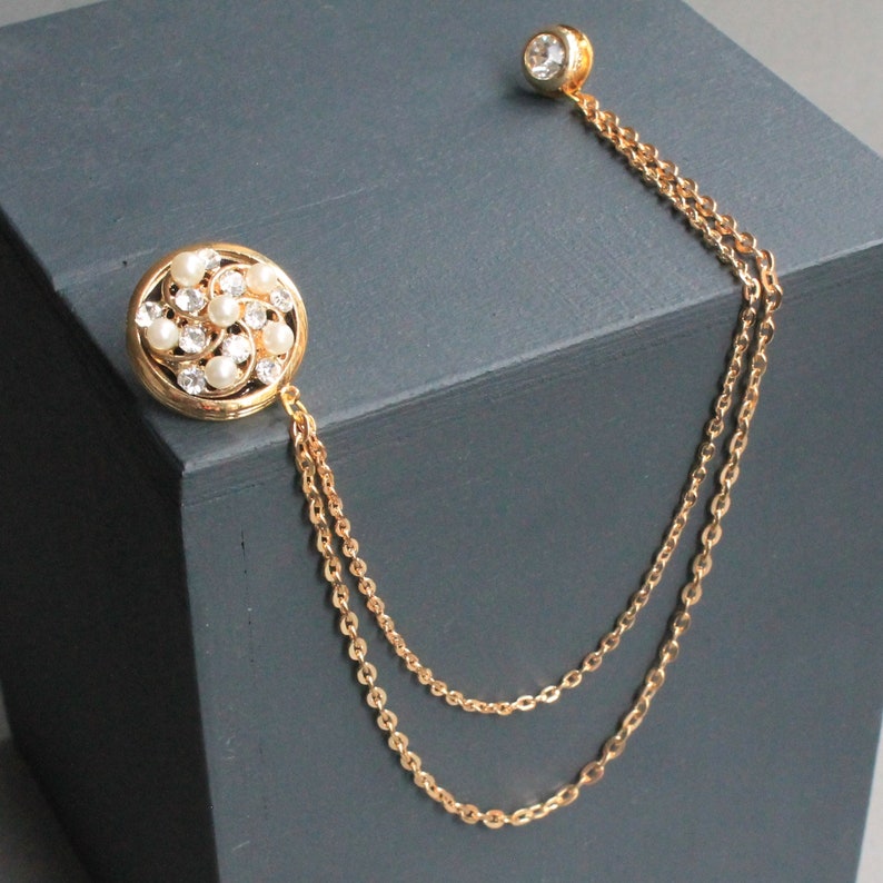 Peut inclure: Une cha&icirc;ne en or avec une broche ronde en perles et strass. La cha&icirc;ne est &agrave; double brin et comporte un petit strass rond en haut.