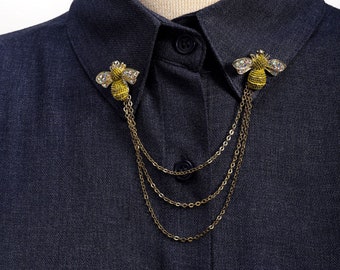 May include: A gold bee brooch with rhinestones, attached to a gold chain, on a dark gray shirt collar.