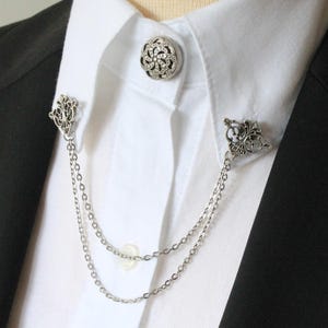 Könnte beinhalten: Eine silberne Kragennadel mit einer dekorativen Kette. Die Nadel hat ein florales Design und ist an einer doppelten Kette befestigt.