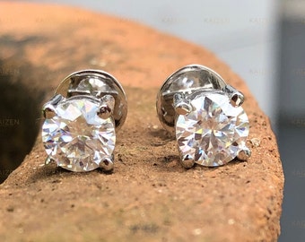 May include: A pair of silver stud earrings with clear round gemstones. The earrings are set on a brown textured surface.