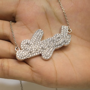 May include: A silver-toned necklace with the name "Love" spelled out in cursive script. The letters are encrusted with small, sparkling crystals. The necklace is displayed on a light-toned background.