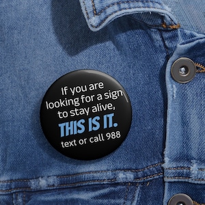 May include: A black button with white and blue text that reads: "If you are looking for a sign to stay alive, THIS IS IT. text or call 988." The button is set against a blue denim jacket.