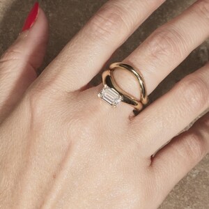 May include: A gold ring with a rectangular, clear gemstone is worn on a hand with red nail polish. The ring has a unique, open design with a curved band. The background is a neutral, textured surface.