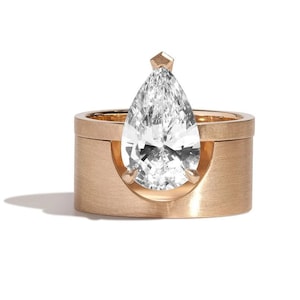 May include: A gold ring with a large, pear-shaped diamond. The diamond is set in a unique design, with a small, geometric accent at the top. The ring has a brushed finish and a modern aesthetic.