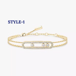 May include: A gold chain bracelet featuring a horizontal oval-shaped charm encrusted with small clear stones and three larger round stones set inside. The text STYLE-1 is visible in the upper left corner, and a small tag reads MESSIKA.