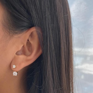 Puede incluir: Primer plano de una oreja con dos pendientes de oro. Los pendientes tienen una gema cuadrada y transparente. El pendiente superior es más pequeño que el inferior. Los pendientes están engastados en oro.