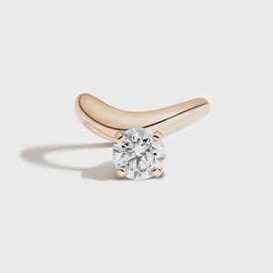 May include: A single, gold-colored earring with a curved design. The earring features a round, clear diamond at its center, held in place by four small prongs. The earring is set against a white background.
