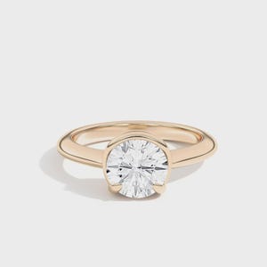 May include: A gold ring with a large, round, clear gemstone. The ring has a simple, elegant design with a smooth band and a bezel setting that holds the gemstone securely. The ring is set against a white background.