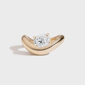 Könnte beinhalten: Ein goldener Ring mit einem großen, klaren Diamanten im Kissenschliff. Das Band des Rings hat ein glattes, geschwungenes Design. Der Diamant wird von vier Krappen gehalten. Der Ring ist vor weißem Hintergrund.