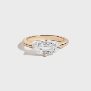 May include: A gold ring with a large, clear, rectangular-cut gemstone. The ring has a simple, polished band and four prongs securing the gemstone. The setting is a warm, yellow gold color, and the gemstone sparkles brightly.