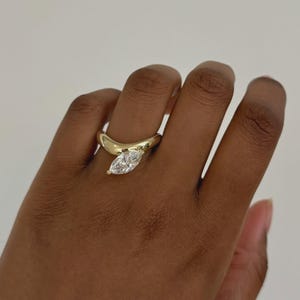Könnte beinhalten: Ein goldener Ring mit einem Diamanten im Marquiseschliff. Der Ring hat ein einzigartiges, geschwungenes Design. Der Diamant ist in einer Krappenfassung gefasst. Der Ring befindet sich an einem Finger. Der Hintergrund ist einfarbig weiß.