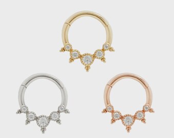 May include: Three hinged nose rings in gold, silver, and rose gold. Each ring features a row of clear, round gemstones. The gemstones are set in a decorative, beaded design. The rings are displayed against a neutral background.
