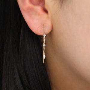 Puede incluir: Un pendiente de color dorado con un delicado diseño de cadena. El pendiente presenta varias piedras preciosas pequeñas y brillantes y un elemento de barra lineal. El pendiente se lleva en la oreja, mostrando su estilo elegante y minimalista.