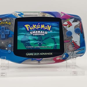 May include: A blue and clear Game Boy Advance handheld console featuring a custom Pokémon Emerald Version theme. The screen displays the Pokémon Emerald Version title logo, and the casing is decorated with graphics of Latias and Latios.